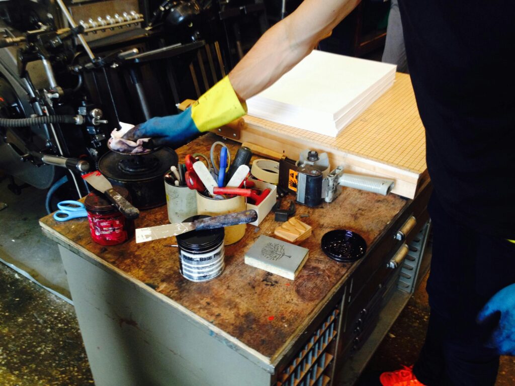Printshop workspace with tools and inks, showcasing a worker's hand in action.